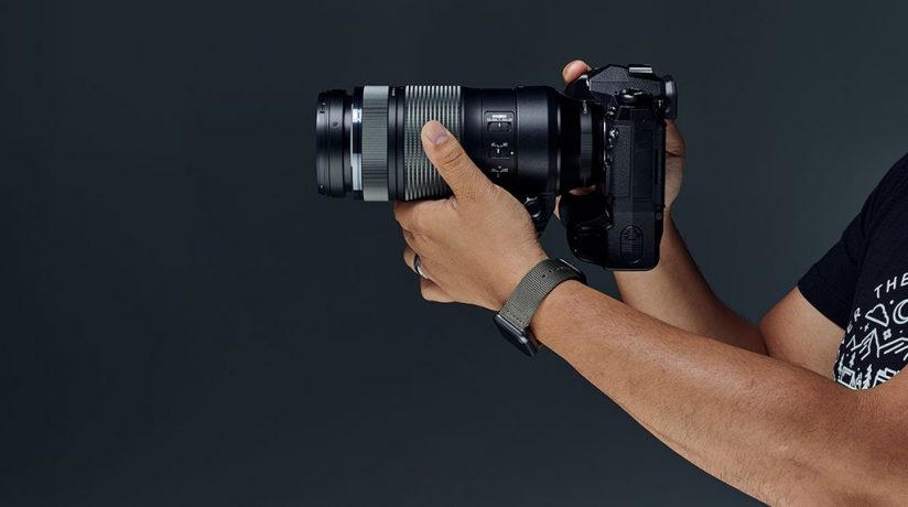 Image of a person holding a camera with the Side view of the Olympus M.Zuiko Digital ED 100-400 f5.0-6.3 IS lens attached