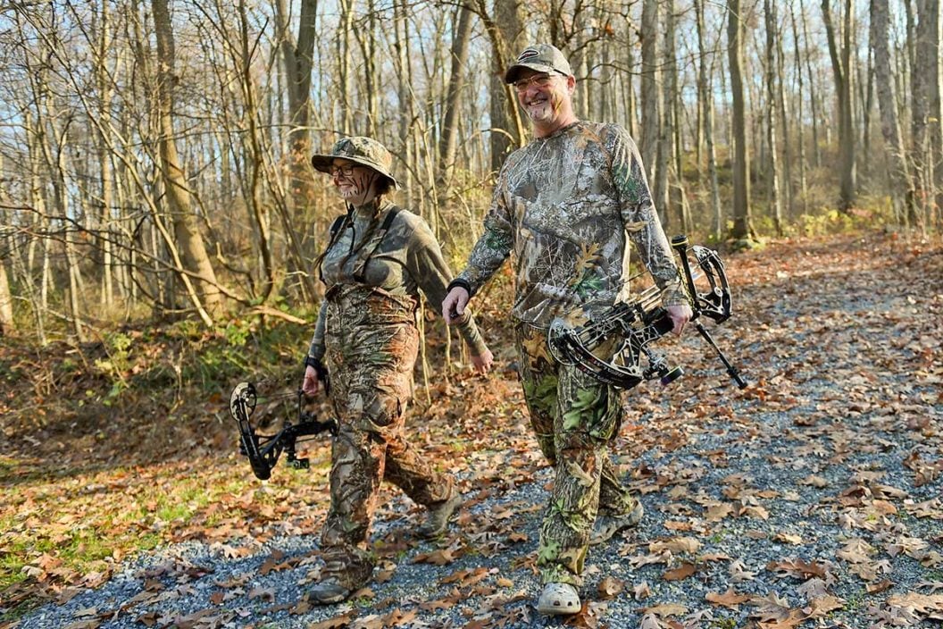 A father and daughter in camo walk in the woods holding hunting bows.