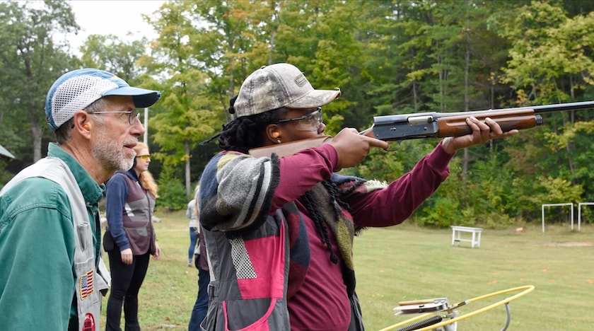an african american woman raises a shotgun at a shooting range. a white male instructor peers over her shoulder
