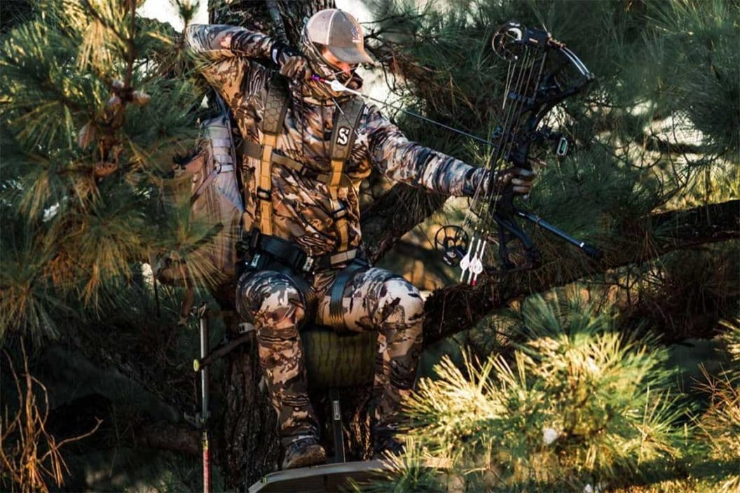 A hunter sits in a lock-on treestand while drawing his bow.